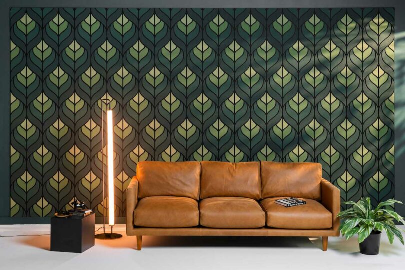 A brown leather sofa is positioned against a wall with green leaf-patterned wallpaper. An illuminated floor lamp, black side table, and potted plant are nearby. A book rests on the sofa