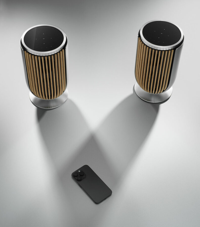 Two cylindrical Beolab 8 speakers with wooden slats and black tops are placed side-by-side on a light surface. In front of them lies a smartphone. Shadows from the speakers converge between the devices.