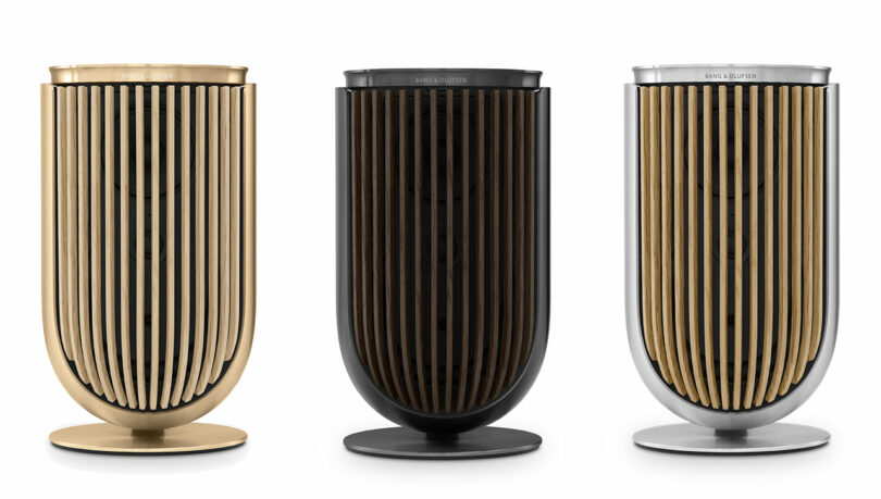 Three Beolab 8 cylindrical speakers with vertical slats, available in gold, black, and silver finishes, are aligned side-by-side against a white background.