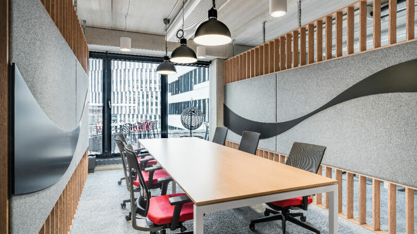 A modern conference room with a long table, red and black chairs reminiscent of Coca-Cola branding, grey acoustic panels, black pendant lights, and large windows overlooking a cityscape.