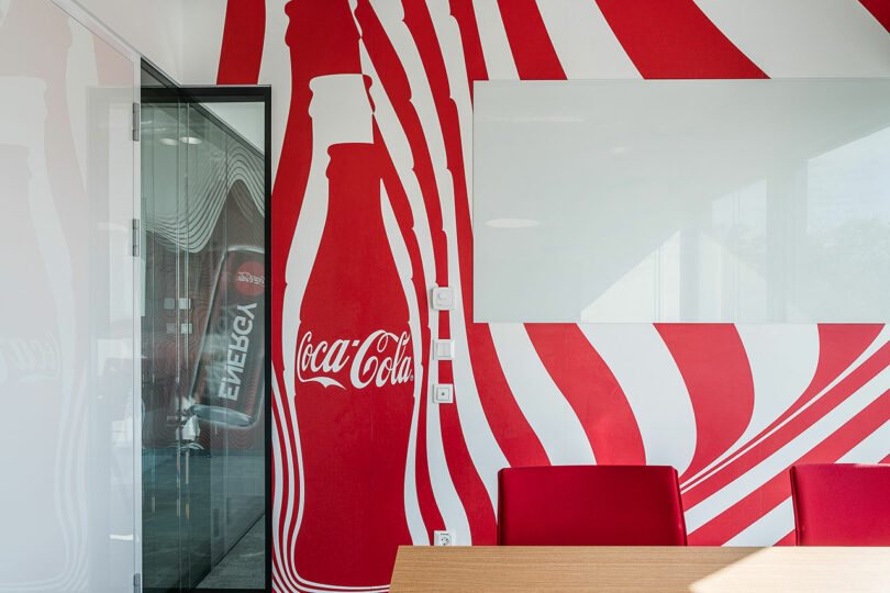 A conference room with glass walls and vibrant red chairs, featuring a striking mural of a large Coca-Cola bottle with white wavy lines on a red background. A whiteboard is mounted on the wall.