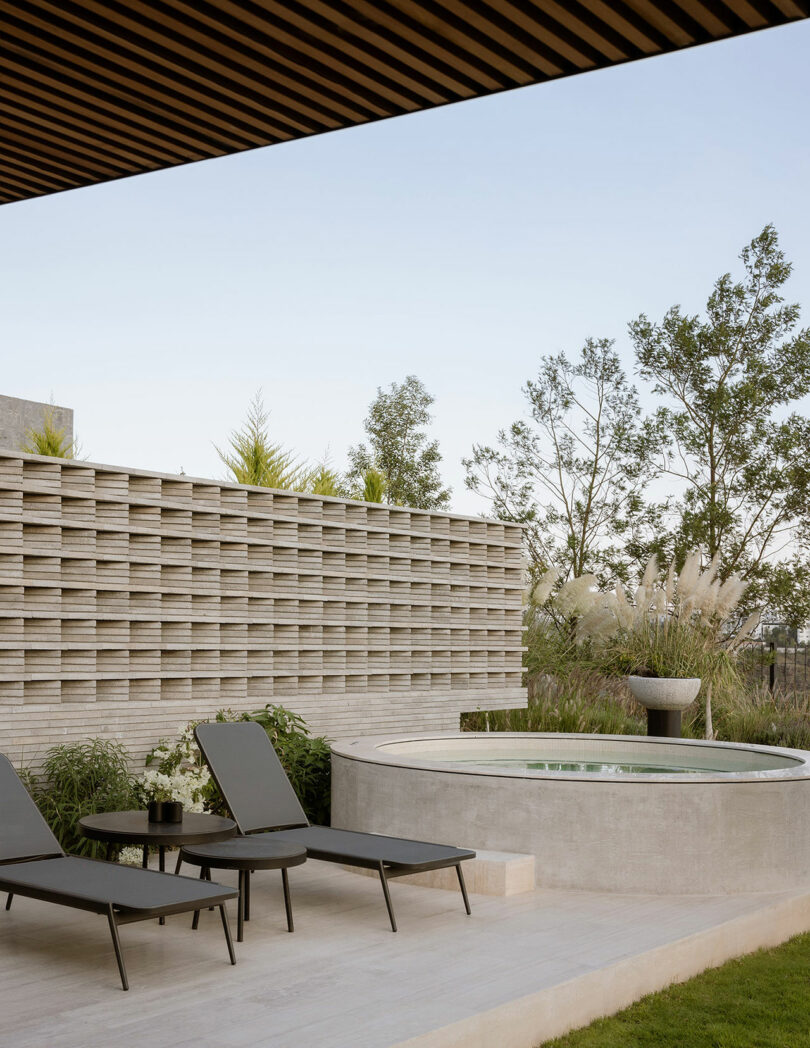 A modern outdoor patio featuring two black lounge chairs with a small table beside a circular hot tub, surrounded by greenery and a textured privacy wall.