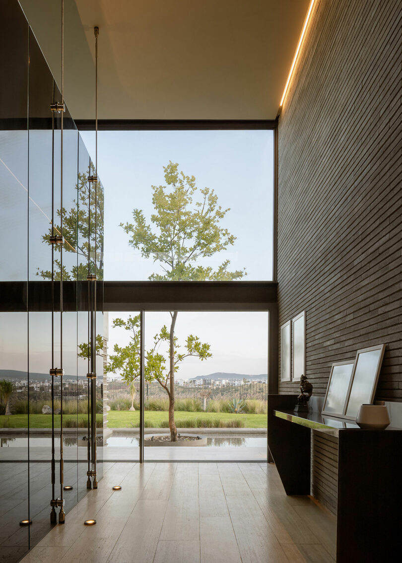 Modern interior with floor-to-ceiling windows showcasing an outdoor tree and landscape view; features minimalist decor, a glass partition, and art pieces on a sleek black console table.