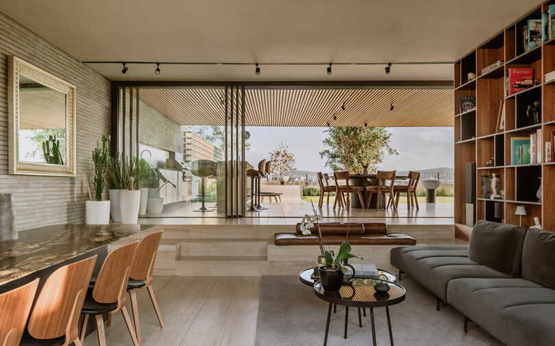 Modern open-concept living space with wooden furniture, large windows, and an outdoor dining area visible through sliding glass doors. A comfortable couch, indoor plants, and a bookshelf are featured.