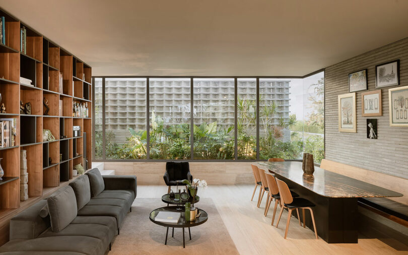 A modern living room features a large sectional sofa, round coffee tables, a spacious dining table with chairs, and a wall of floor-to-ceiling bookshelves, with large windows showcasing an outdoor garden.