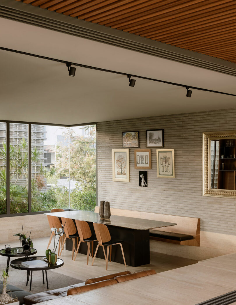 A modern dining area with a long black table, wooden chairs, and framed art on the walls. Large windows reveal outdoor greenery; wooden ceiling panels and indoor plants add to the contemporary decor.