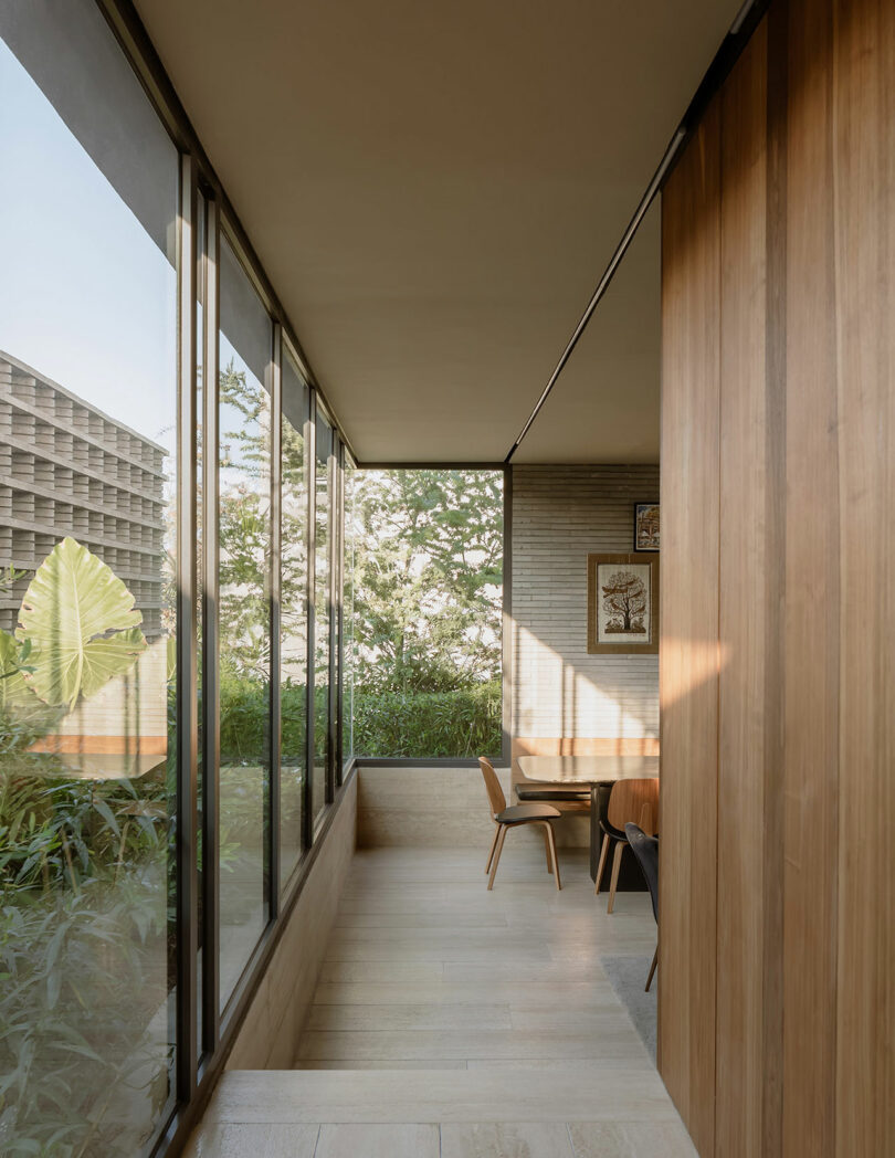 A modern interior space with large glass windows, wooden walls, and floors. There is a small table with chairs near the windows, overlooking green foliage outside.