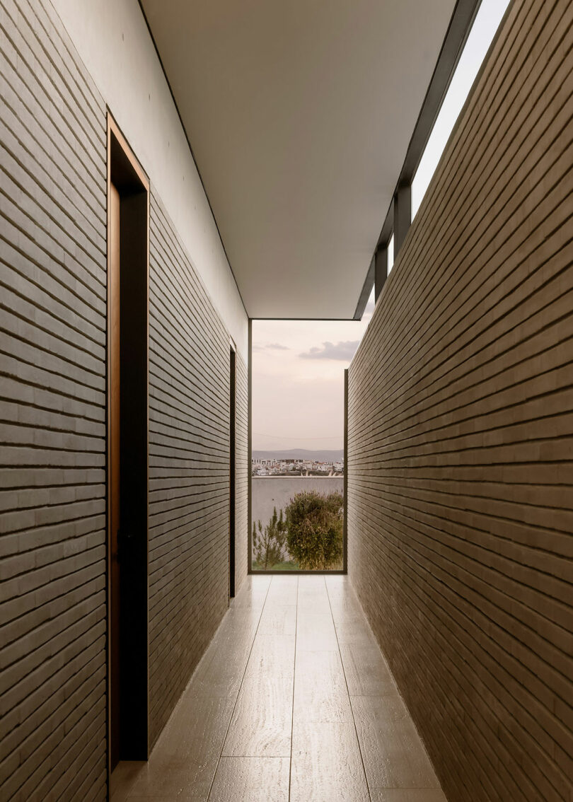 A narrow corridor with brick walls and a large window at the end, offering a view of a cityscape and sky.