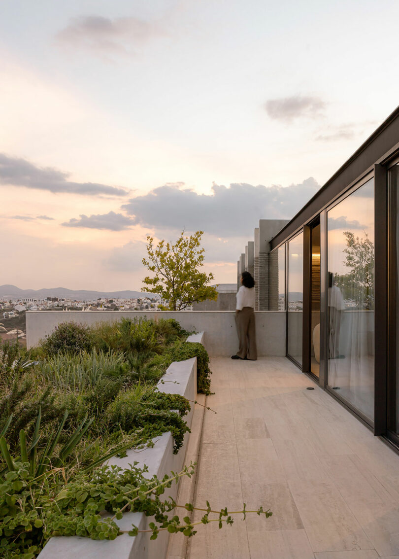 Person standing on a terrace with planters, looking out at a cityscape during sunset.