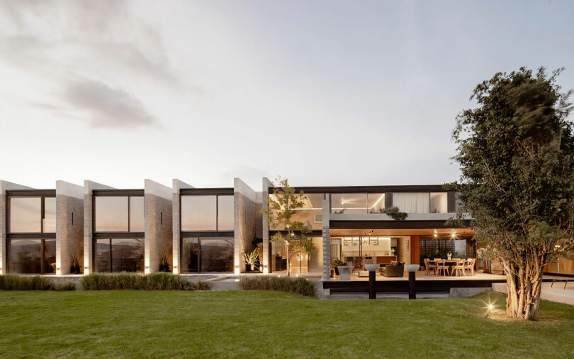 Modern two-story house with large windows, an outdoor dining area, and a spacious lawn in the foreground.