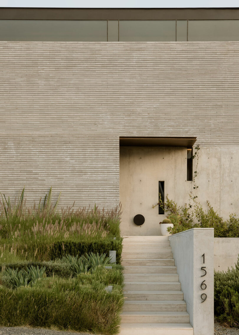 A modern minimalist house with a vertical brick facade, a rectangular door, and a staircase leading to the entrance. The house number "1569" is displayed on the side of the stairway. Lush greenery surrounds the steps.