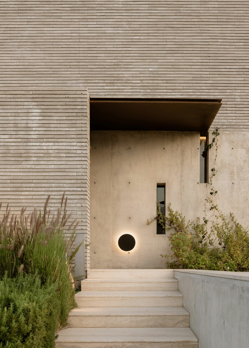 A minimalist entrance with concrete walls, a circular light fixture near the door, and a small rectangular window. The front steps are surrounded by greenery.