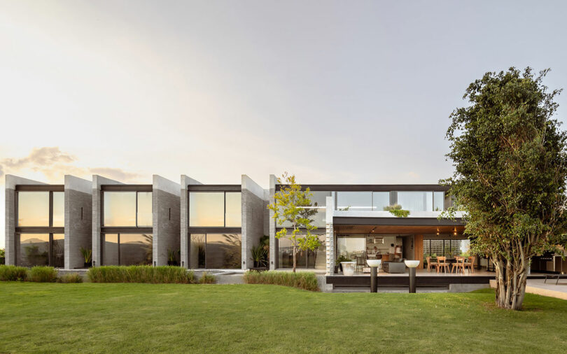 Modern house with large glass windows and concrete design, featuring an open outdoor dining area and well-maintained lawn.