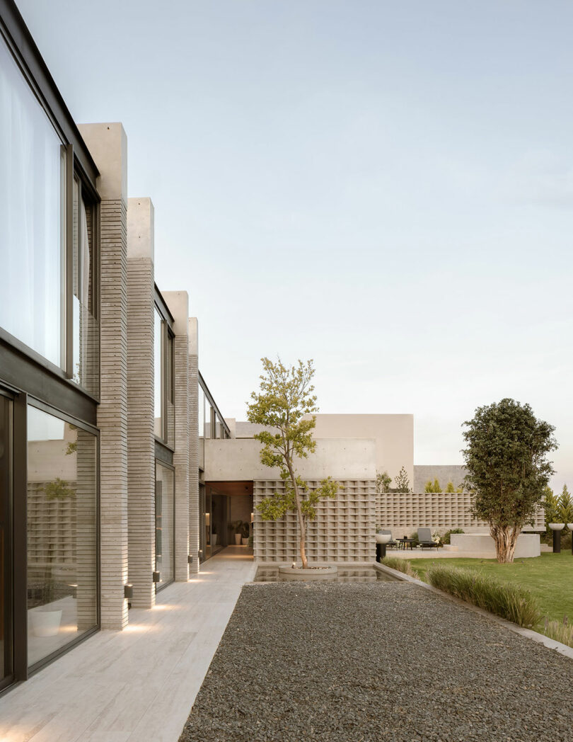 Modern, minimalist house with large windows, concrete walls, and gravel courtyard surrounded by greenery and a few trees. Clear sky in the background.