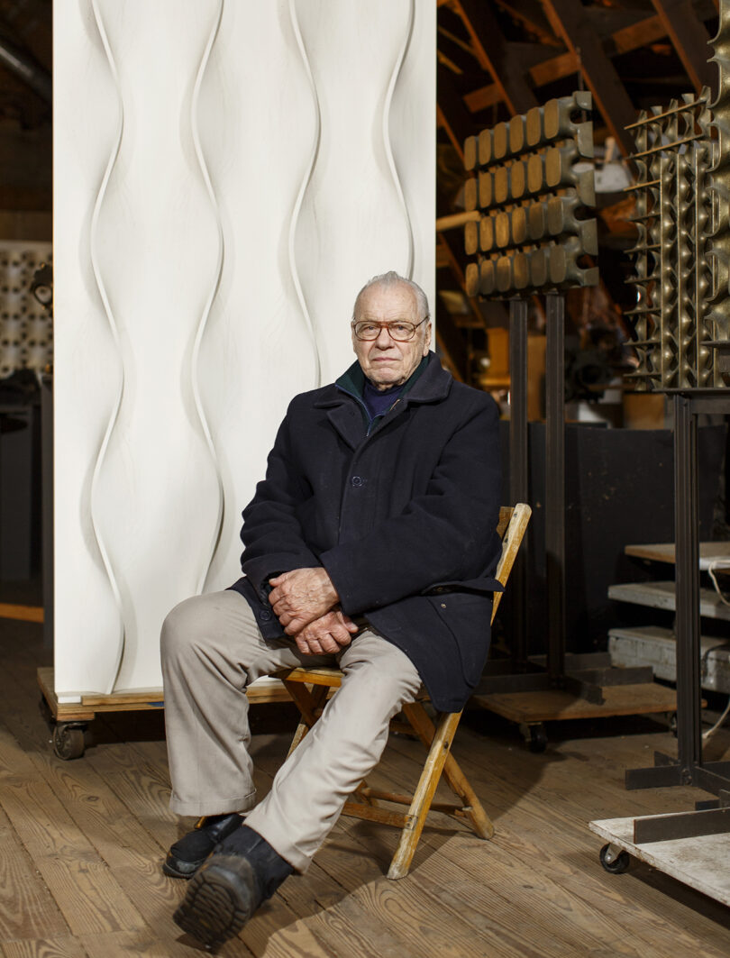 Erwin Hauer wearing glasses and a dark coat sits on a wooden chair in a workshop with large sculptural panels in the background.