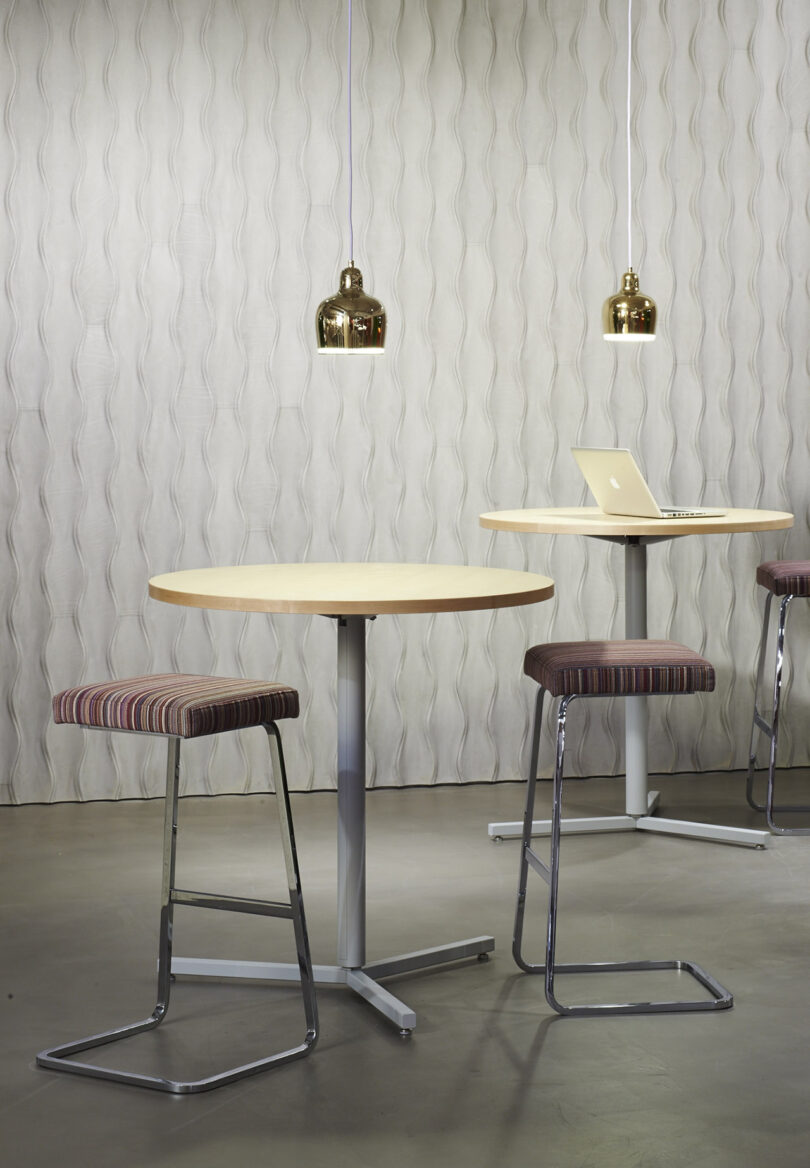 Modern workspace featuring round tables with stools, a laptop on one table, and two hanging pendant lights. The background has a textured, wave-patterned wall, Design 406 by Erwin Hauer.