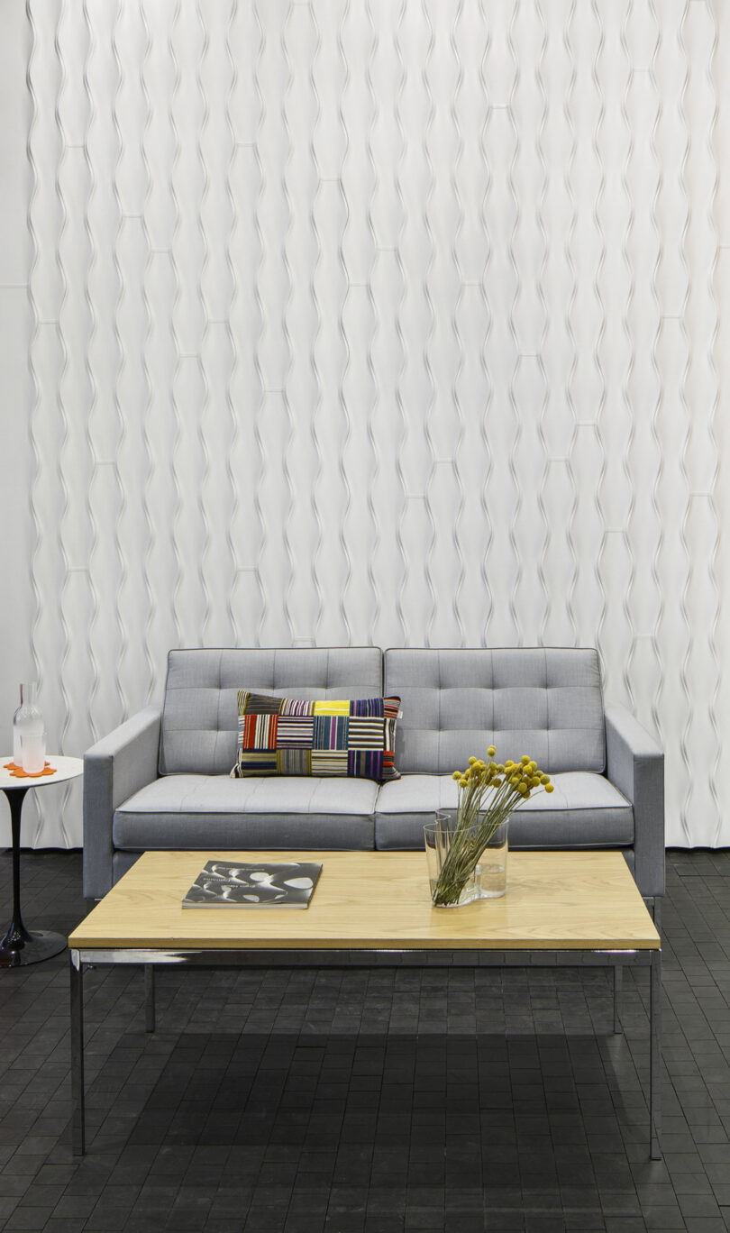 A modern living room with a grey sofa, colorful pillows, a wooden coffee table with a glass vase of yellow flowers, a magazine, and a black side table with a drink and fruit on it. In the background there is a wavy, undulating wall covering, Design 406 by Erwin Hauer.