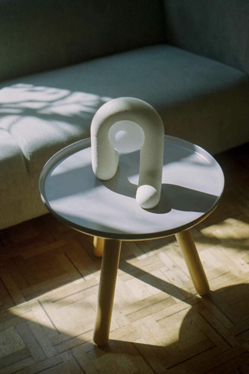 A small sculptural piece with an arch shape sits on a round table with wooden legs, next to a couch. Sunlight casts shadows on the table and floor