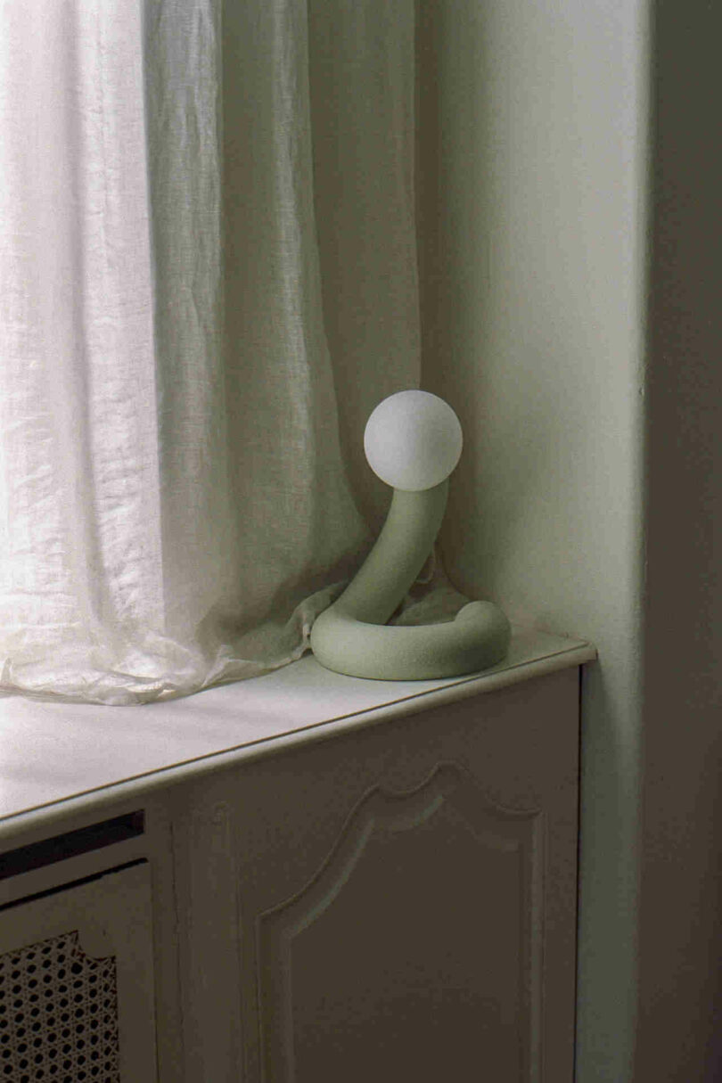 A white-cushioned lamp with a round bulb stands on a windowsill next to a pale curtain