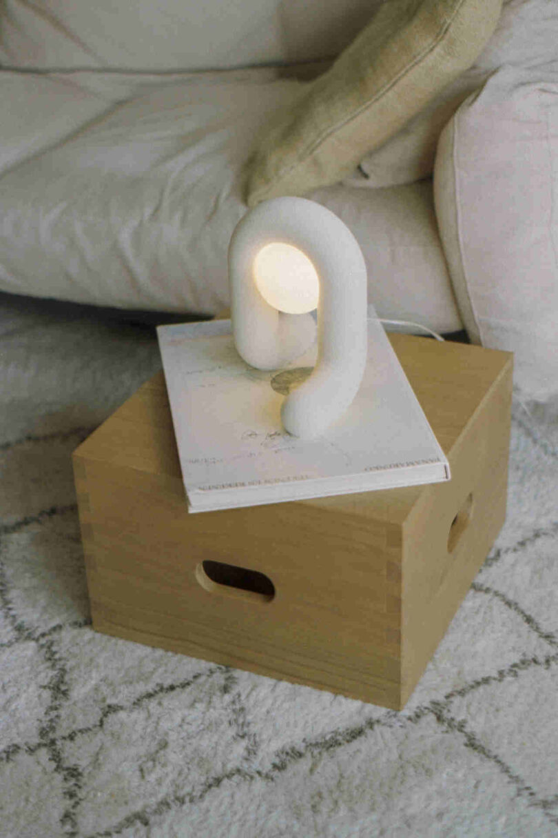 A small white table lamp with a unique arch design is placed on a wooden box beside a couch. The lamp is lit, casting light on an open book beneath it