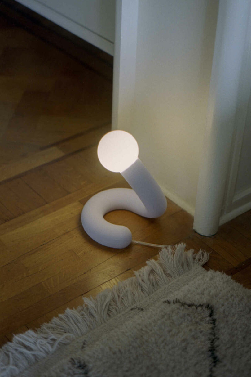 A modern, white floor lamp curves into a question mark shape, with a round light bulb at the top, placed on a wooden floor next to a fringed rug