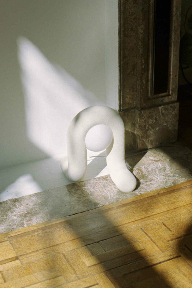 A white sculptural art piece resembling an abstract loop is placed on a wooden floor and partially lit by sunlight