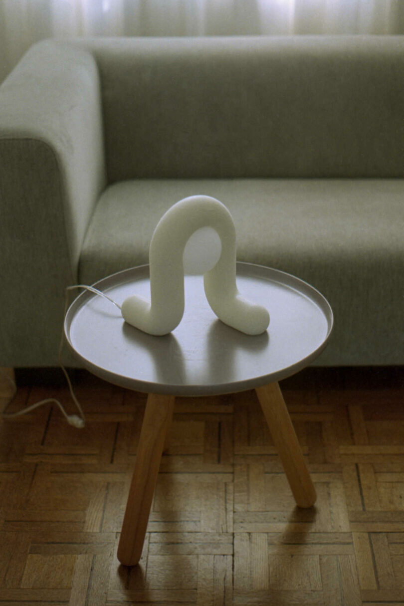 A U-shaped modern lamp sits on a round wooden coffee table in front of a grey couch, with a sheer curtain in the background