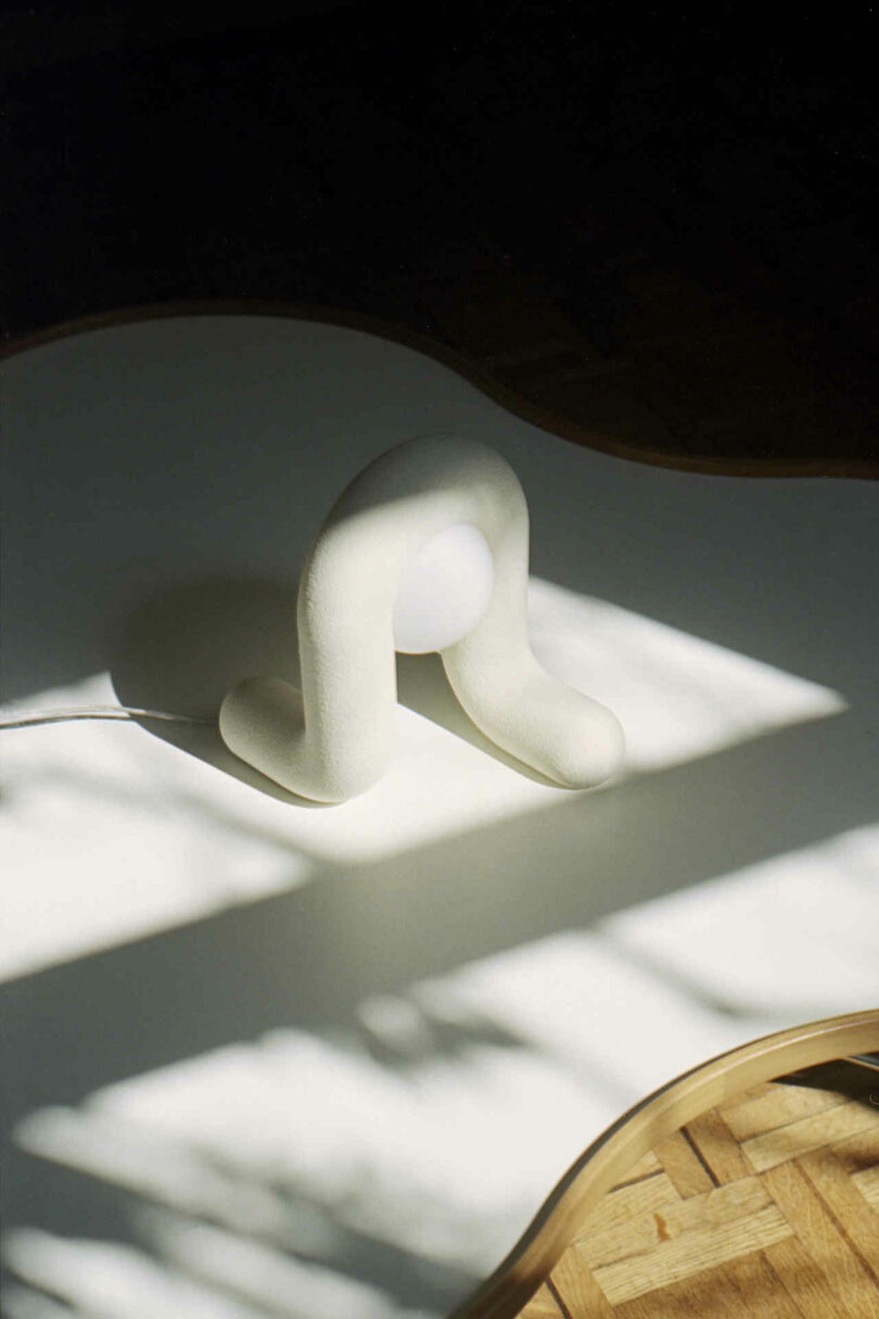 A curved, cream-colored object with two bent legs and a round white sphere in the middle, placed on a white surface with partial sunlight and shadows