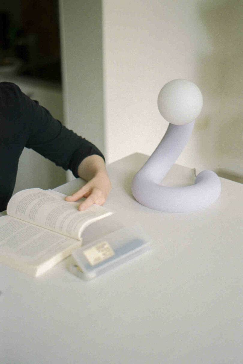 A person in a dark shirt reads a book at a table with a unique white lamp featuring a curved design and a spherical top. A small closed book lies nearby