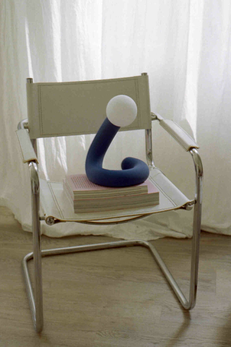 A white chair with metallic armrests holds a stack of books with a blue sculptural figure topped by a white sphere on top, placed in front of a sheer curtain