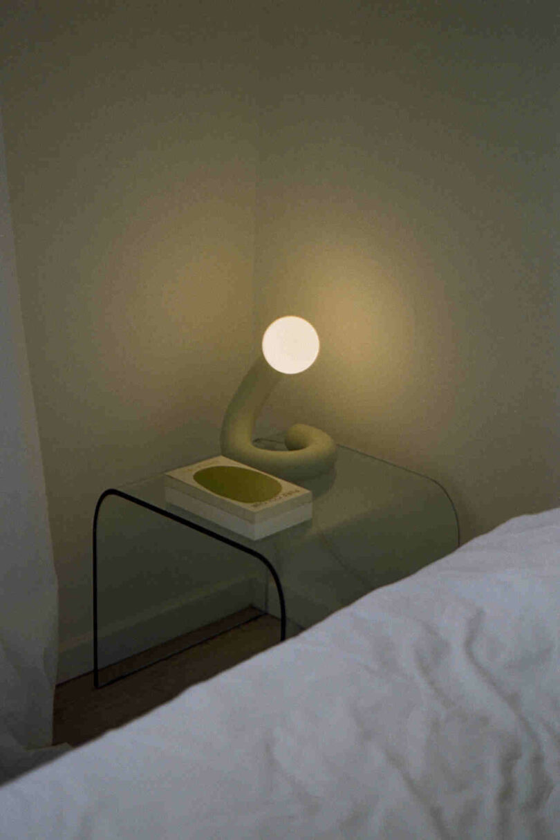 A modern bedside table lamp with a round bulb and a curved base is lit beside a bed with a white sheet. A white rectangular tray is placed on the transparent glass table