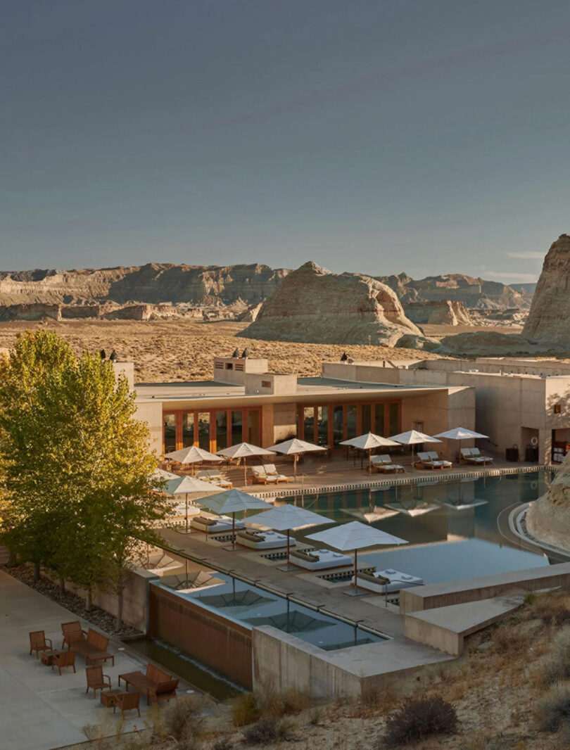 A luxurious desert resort with a pool, lounge chairs, and umbrellas, set against a backdrop of rocky desert formations and clear skies.