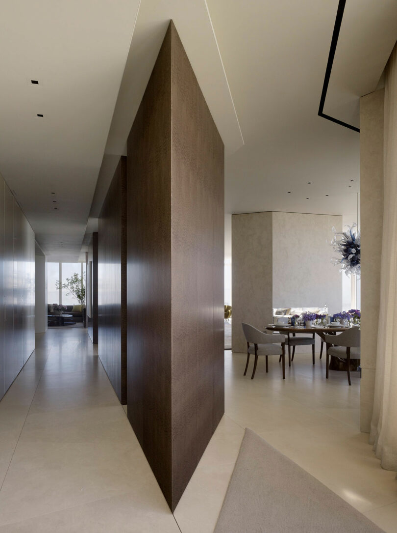 Modern interior with a long corridor featuring sleek walls and recessed lighting. A dining area with a set table and contemporary chairs is visible to the right, and a seating area is in the background.