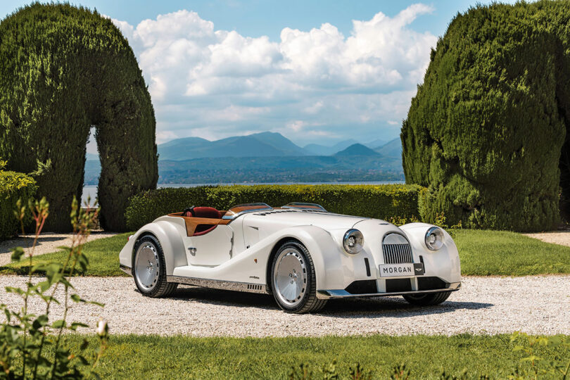 A white Morgan roadster, designed by Felix Kilbertus, is parked on a gravel path surrounded by lush greenery and hedges, with mountains visible in the background under a partly cloudy sky.