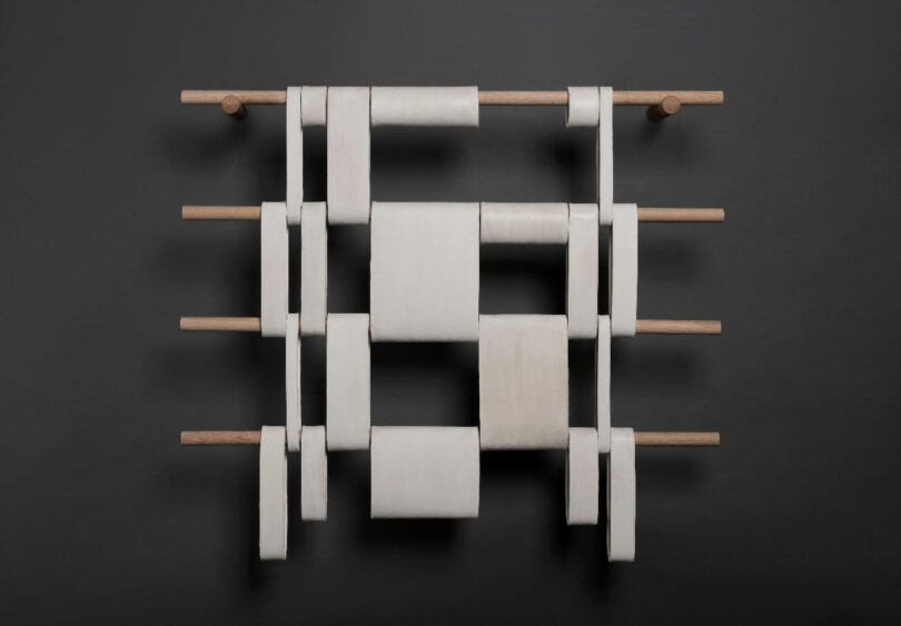 A minimalist sculpture with a grid of intertwined white ceramic or plastic loops held by wooden rods on a dark background.