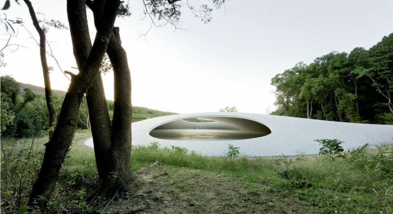 A white, dome-shaped structure with a large circular opening in the center is set amidst a verdant landscape with trees and grass.