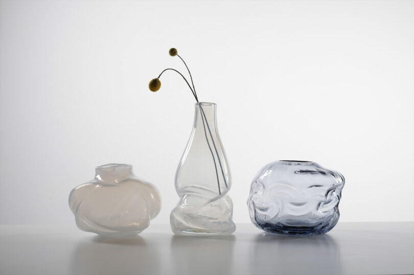 Three artistically shaped glass vases of different transparencies and colors sit on a white surface. The middle vase holds two dried flower stems with round yellow buds.