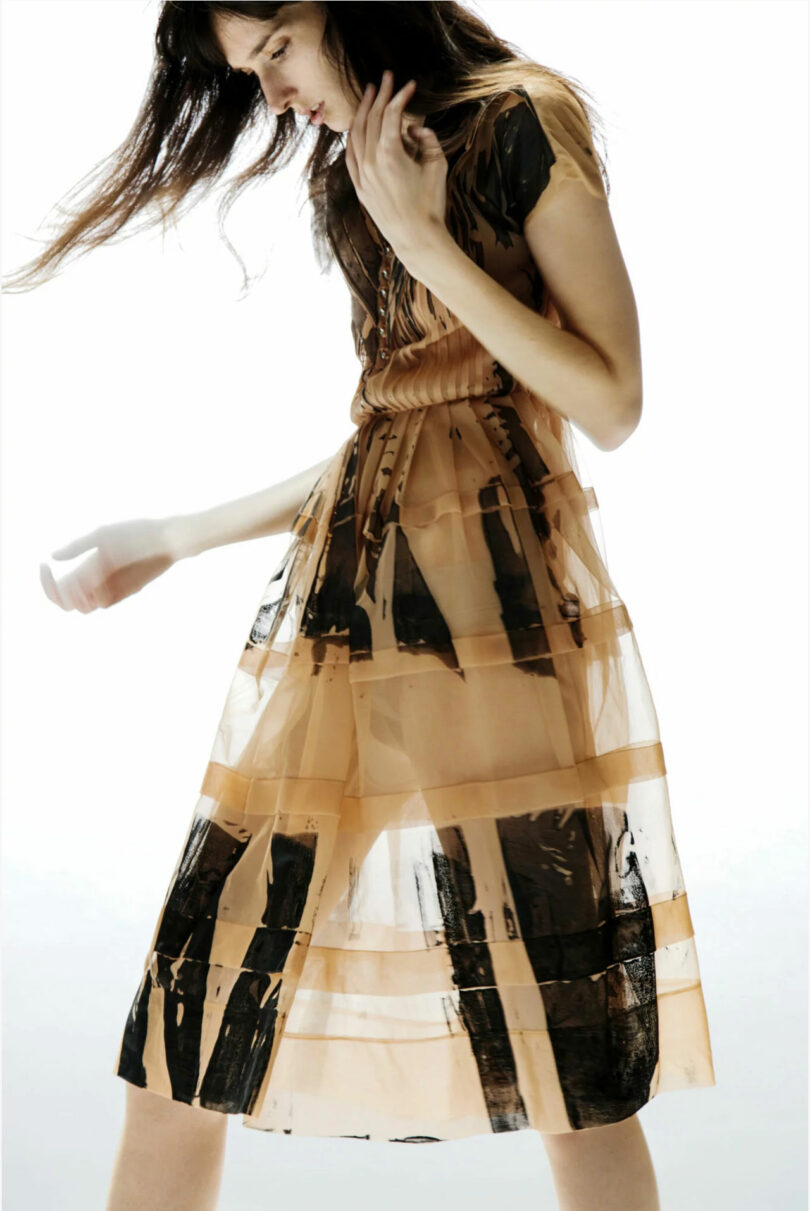 A woman with long hair wearing a semi-transparent, orange and black patterned dress, standing against a white background. She is slightly leaning forward with her left arm raised.