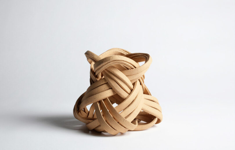 A complex, intertwined, light brown sculptural piece made of what appears to be wooden strips, set against a plain white background.