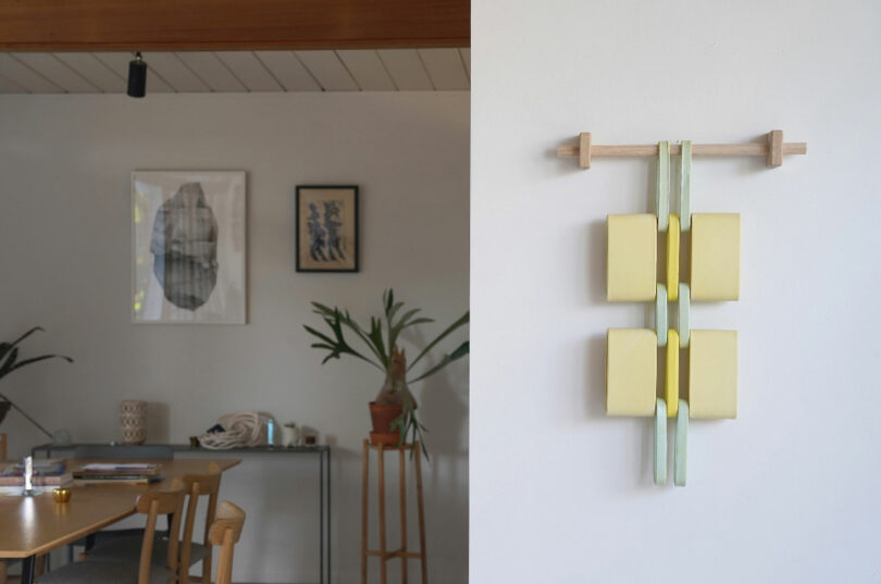 A modern art piece with yellow and green elements is mounted on a white wall. In the background, there is a wooden table, chairs, small plants, and framed artwork on a light-colored wall.