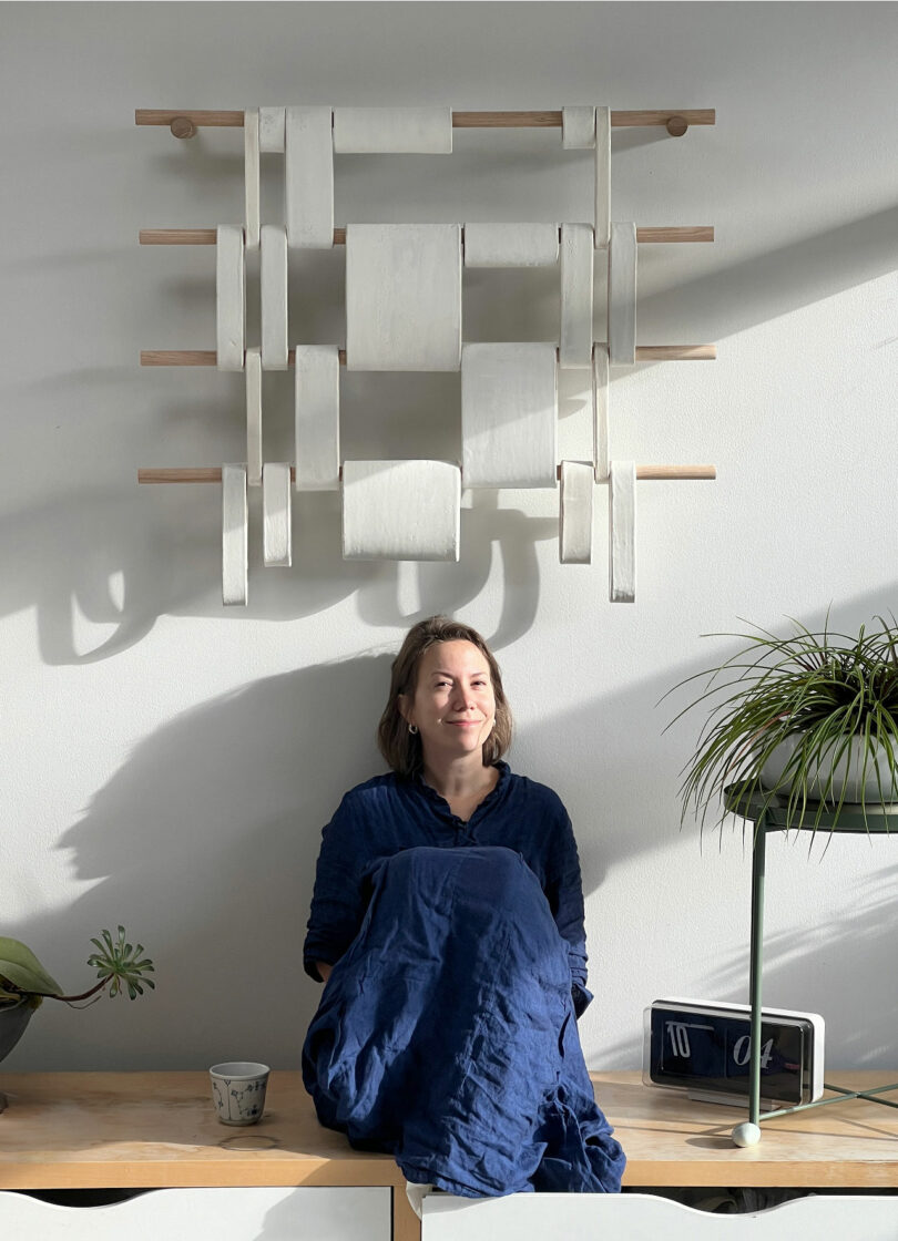 A person sits on a wooden bench beneath a modern wall art piece, surrounded by plants and a clock, dressed in a blue garment while smiling with sunlight illuminating the scene.