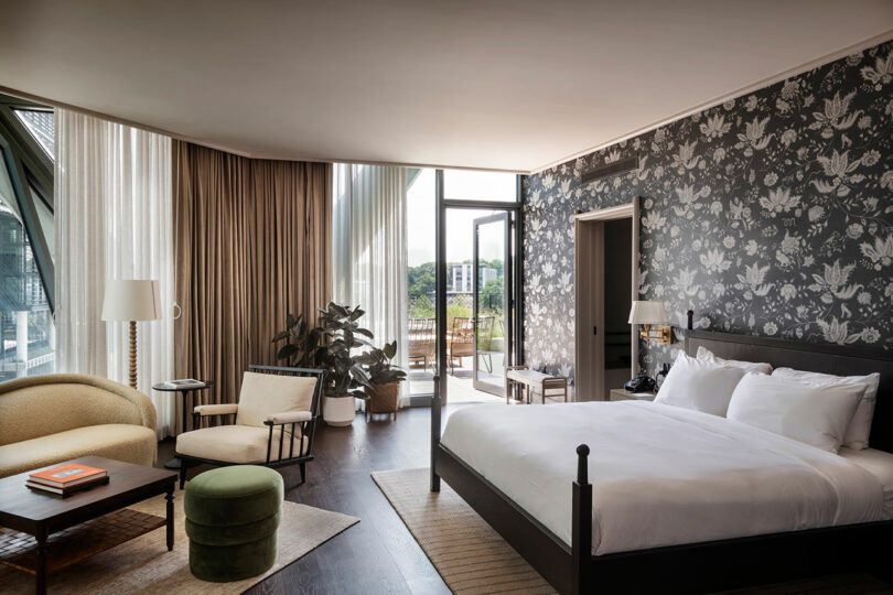 A modern FORTH Hotel room with a king-sized bed, patterned wallpaper, seating area, large window, and a balcony overlooking greenery.