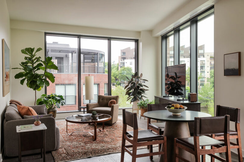 A modern, well-lit living room at FORTH Hotel features a sofa, armchair, coffee table, dining set, plants, and large windows with a city view.