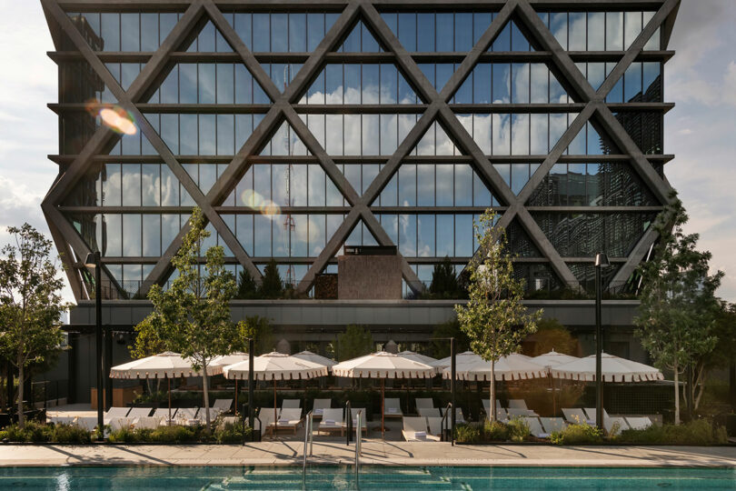 The FORTH Hotel is a modern glass building with a geometric façade, behind which is a pool area with white loungers and parasols. Trees and greenery surround this tranquil oasis.