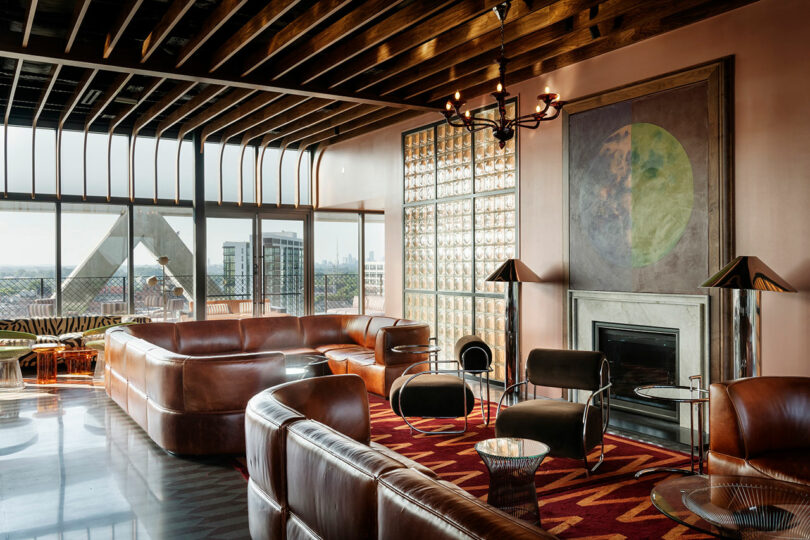 A modern living room in the FORTH Hotel boasts leather sofas, a fireplace, large windows, and artwork on the wall. The ceiling features exposed beams while the floor displays a patterned rug.