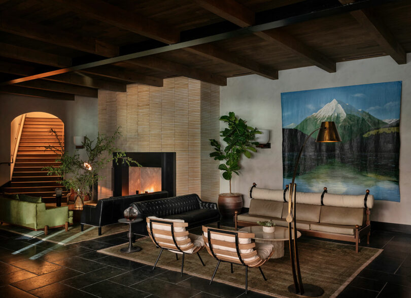 Modern living room with slate flooring at the FORTH Hotel, featuring a black leather sofa, wooden armchairs, and a white cushioned bench. There is a large fireplace, a leafy plant, and a tapestry depicting a mountain.