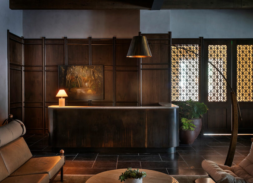 A dimly lit room with dark wood paneling, a dark reception desk, a small lamp, a painting and round tables creates a welcoming atmosphere at the FORTH Hotel. A large plant stands next to the lattice doors and adds a touch of green to the sophisticated ambience.