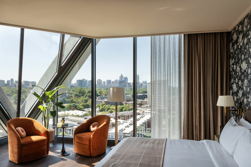 A hotel room at FORTH Hotel with large corner windows offering a city view, featuring a bed, two orange chairs around a small table, a lamp, and a plant. Light flows through sheer curtains.