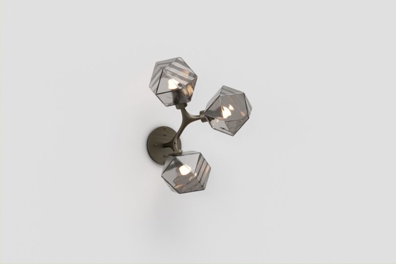 A modern wall sconce with three geometric, multi-faceted glass shades attached to a metal frame, mounted on a plain white wall