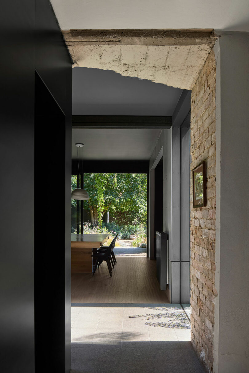 View through a doorway with partially exposed brick and concrete leading to a modern dining area with a wooden table, chairs, and large window showcasing green trees outside.